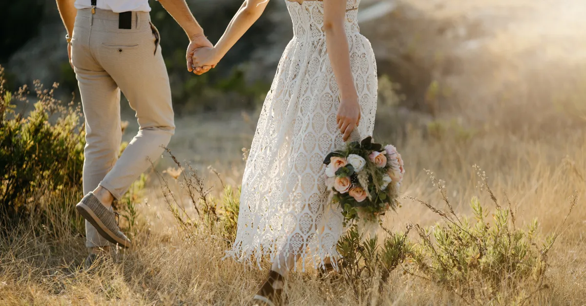 bride and groom holding hands walking through grassy hillside at goldenhour