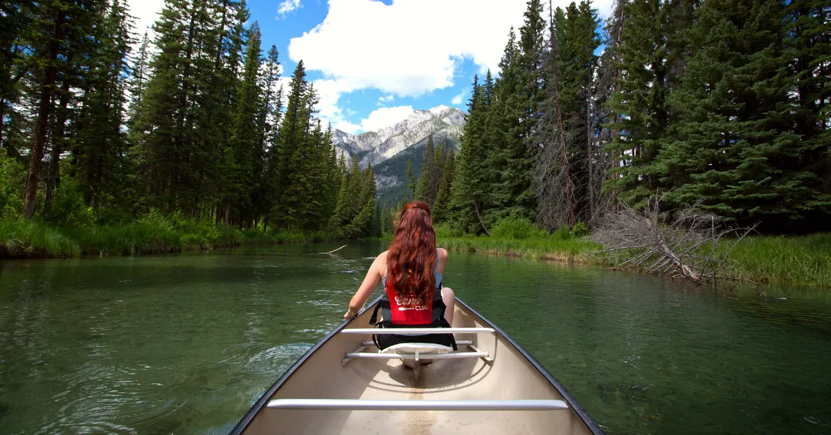 girl in metal canoe in river with pine trees and mountains in background